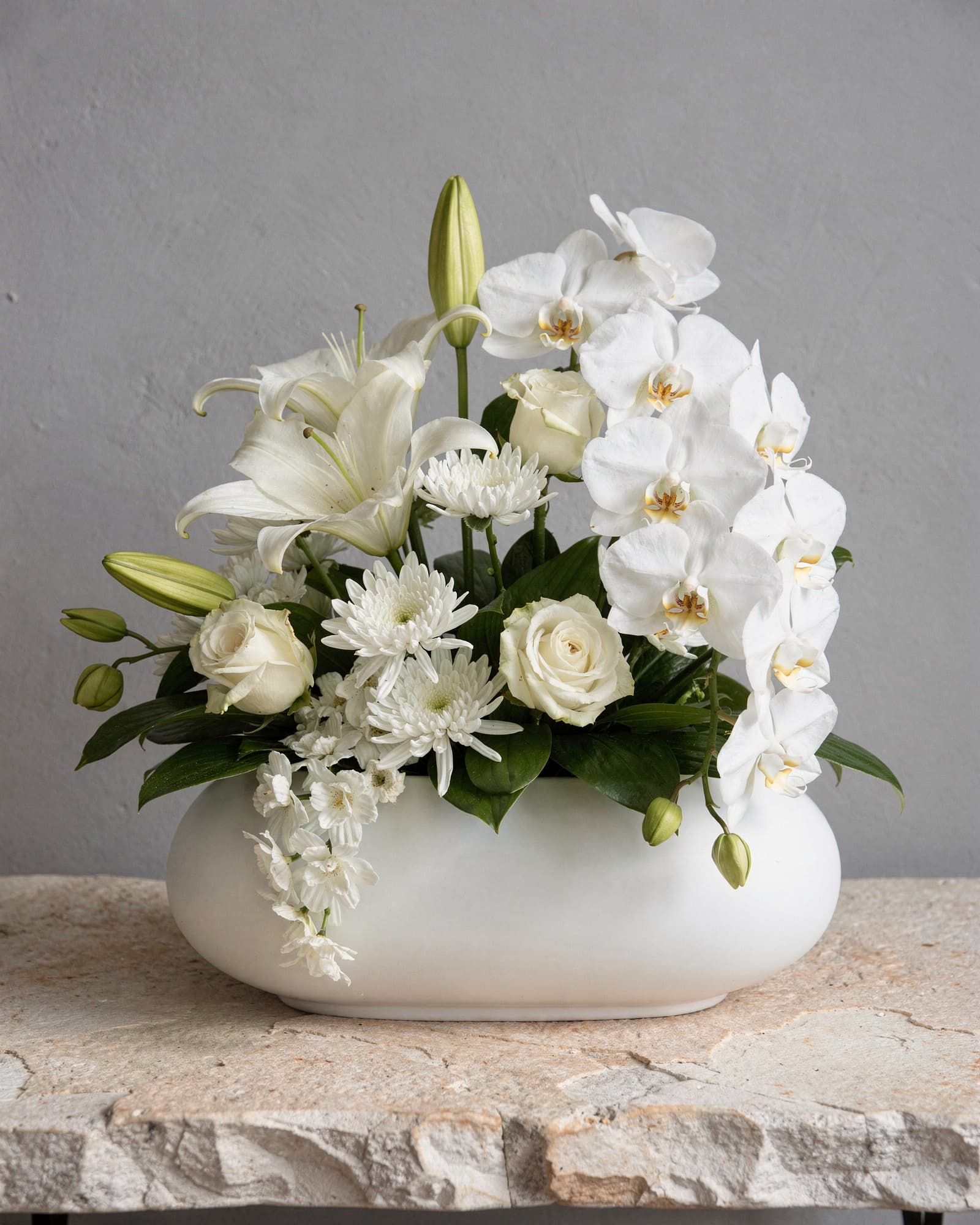 A white arrangement of Phalaenopsis orchids, lilies, roses and chrysanthemums in a low white container