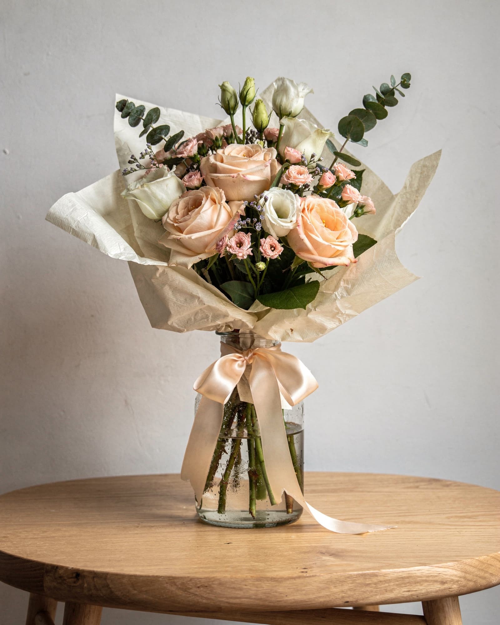 A hand-tied bouquet of peach roses, white lisianthus and eucalyptus wrapped in cream paper, in a glass jar